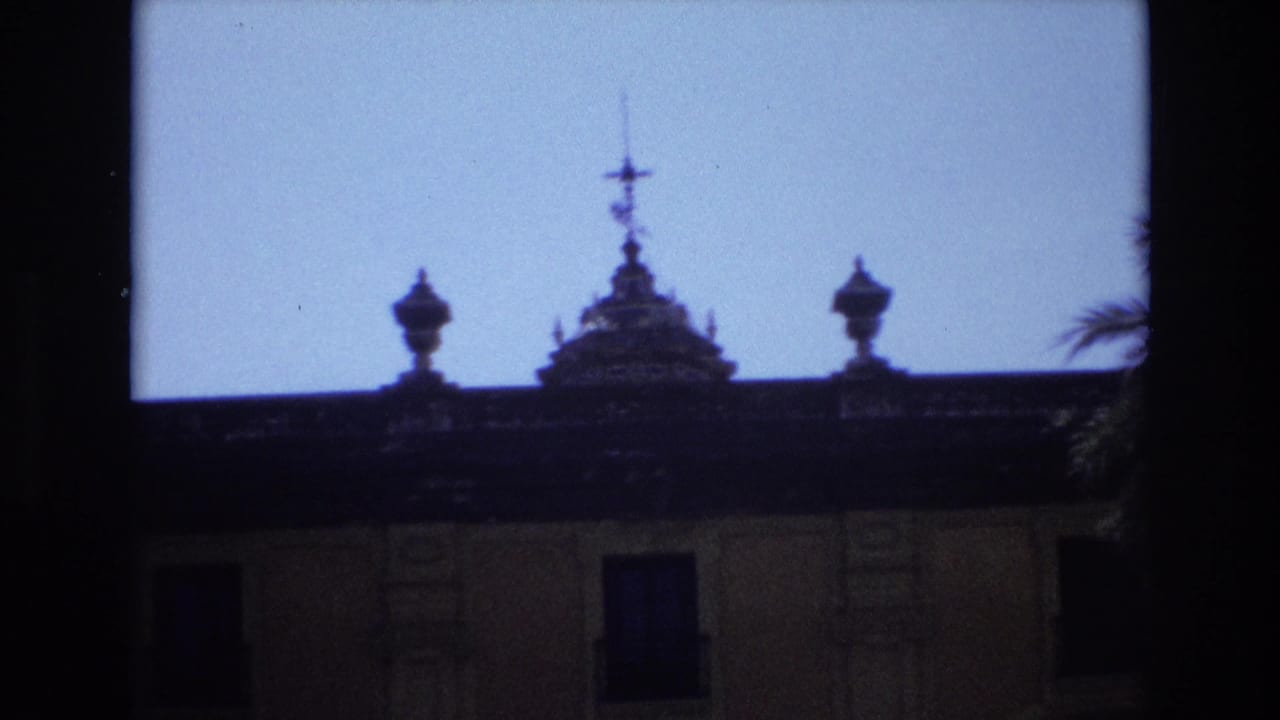 1972 Spain 8mm frame showing the dark roofline, decorative finials, and upper facade of an ornate historic-looking building against a bright sky.