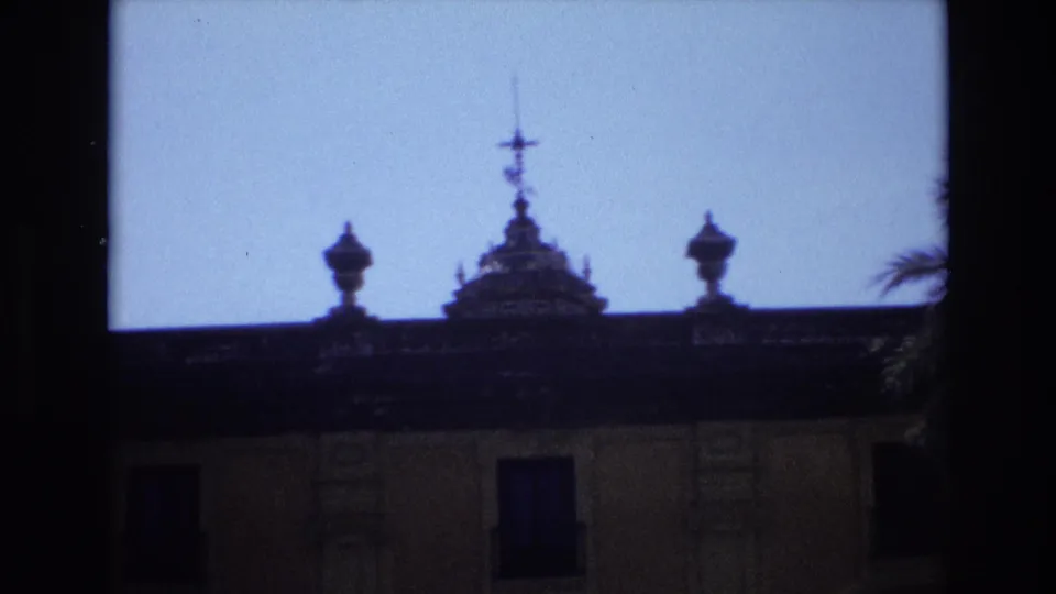 1972 Spain 8mm frame showing the dark roofline, decorative finials, and upper facade of an ornate historic-looking building against a bright sky.