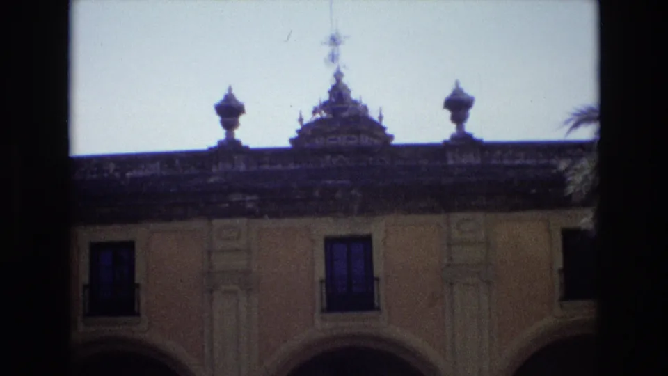 1972 Spain 8mm frame with the camera slightly lower on the pale facade, showing roof ornament, balcony windows, and decorative vertical facade details.