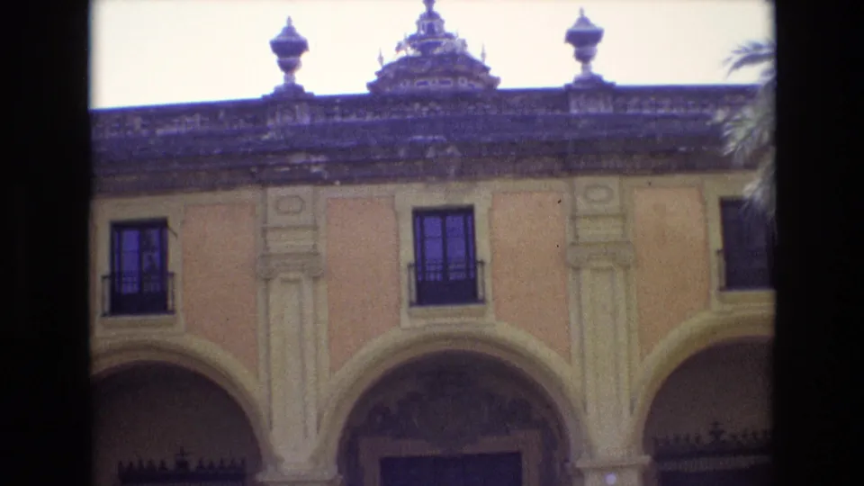 1972 Spain 8mm frame revealing more of the historic-looking facade, including arched entryways, columns, dark balcony windows, and a hint of palm fronds at the side.