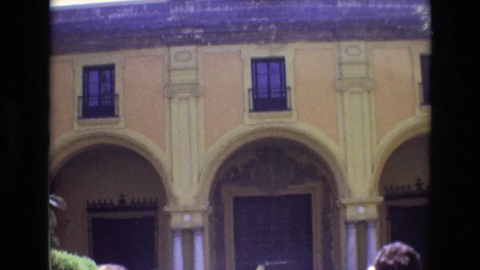 1972 Spain 8mm frame showing the lower arched entrance of the ornate facade, iron gates, decorative columns, and partially visible visitors near the bottom edge.