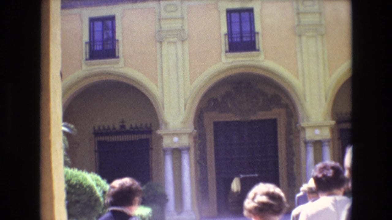 1972 Spain home-movie frame showing tourists gathered in front of a yellow stucco facade with arched openings, columns, and dark upper windows.