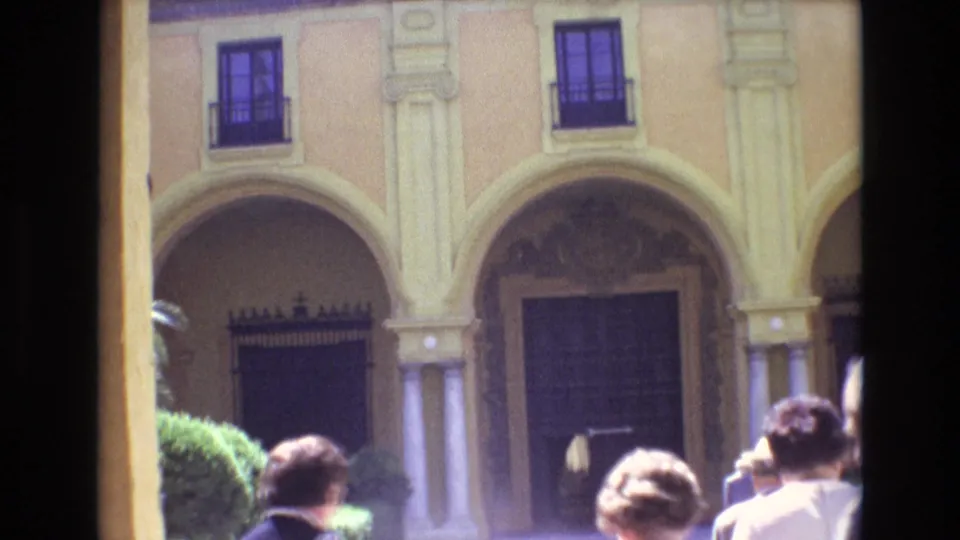 1972 Spain home-movie frame showing tourists gathered in front of a yellow stucco facade with arched openings, columns, and dark upper windows.
