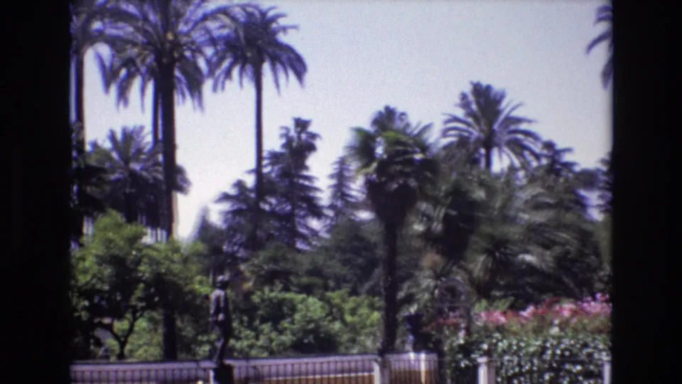 1972 Spain home-movie frame of a cultivated garden with palms, flowering shrubs, a low fence, and a statue seen from a visitor's viewpoint.