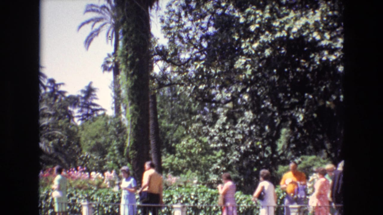 A group of visitors walks through a Spanish garden setting in 1972, with palms, shrubs, flowers, and home-movie frame borders visible.
