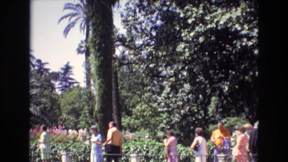 A group of visitors walks through a Spanish garden setting in 1972, with palms, shrubs, flowers, and home-movie frame borders visible.