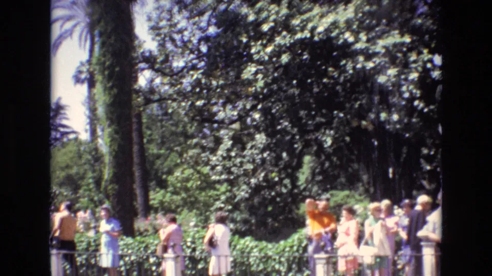 Tourists in period warm-weather clothing gather and walk near a garden railing in Spain, with palms and dense greenery filling the frame.