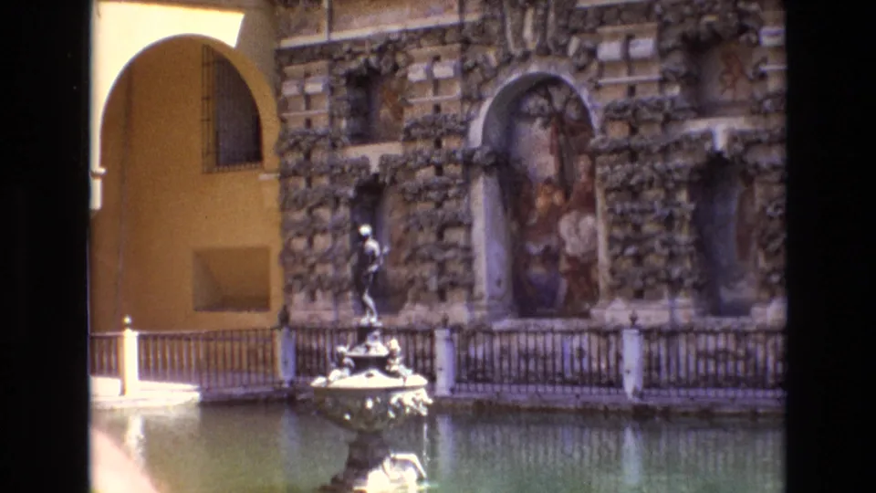 A historic courtyard in Spain shows a stone fountain, water basin, arched doorway, and ornate carved wall details in 8mm home-movie footage.