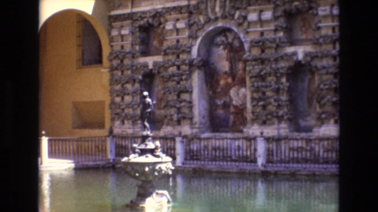 1972 Spain 8mm travel footage showing a sculptural fountain in a courtyard, set against an ornate stone wall with arched niches, painted recesses, railings, and a yellow archway.