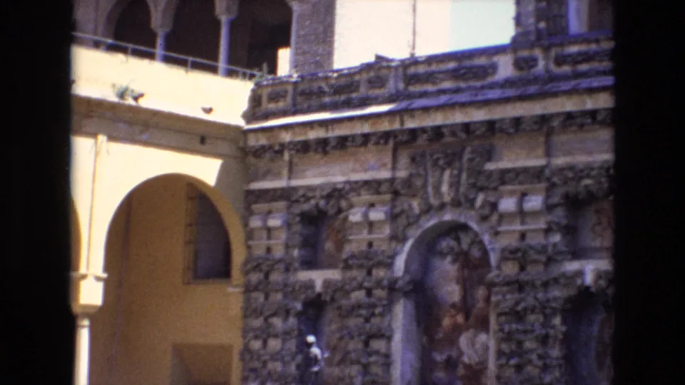 1972 Spain 8mm frame showing the fountain and statue from a slightly lower angle, with textured stonework, a painted niche, railings, and a yellow arched passage in the courtyard.