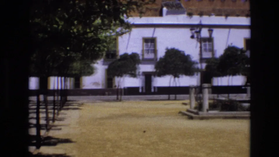 1972 Spain travel footage showing a quiet paved courtyard with trees, benches, lamp posts, a white building facade, windows, and shaded walkways.