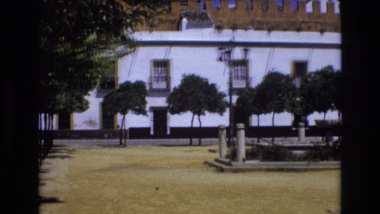 1972 Spain home-movie frame showing a sunlit square with a white building, crenellated roofline, trimmed trees, and a fountain area in the foreground.