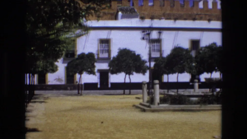 1972 Spain home-movie frame showing a sunlit square with a white building, crenellated roofline, trimmed trees, and a fountain area in the foreground.