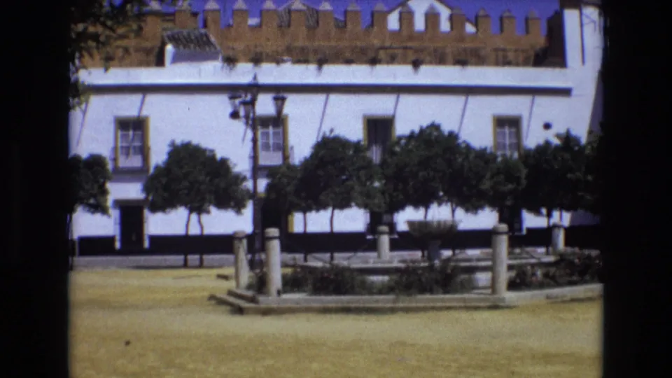 1972 Spain frame of a quiet public square with a white building behind trimmed trees and a circular fountain bordered by low stone posts.