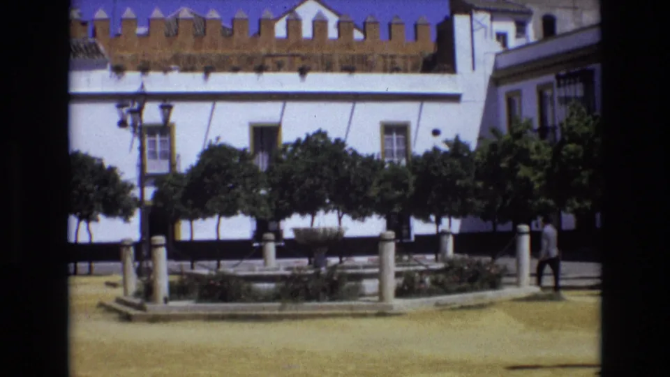 1972 Spain home-movie frame showing the square during the pan, with a fountain, low posts, trimmed trees, and a distant man walking at the right edge.