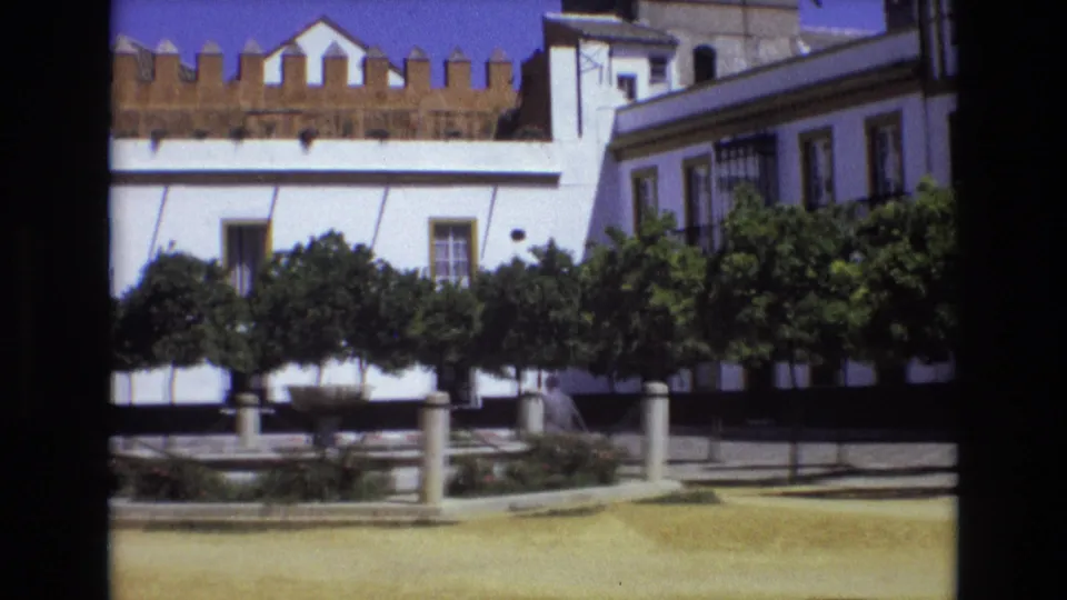 1972 Spain frame near the end of the pan, showing a white building, fountain, trees, and a distant person crossing or standing near the right side of the square.