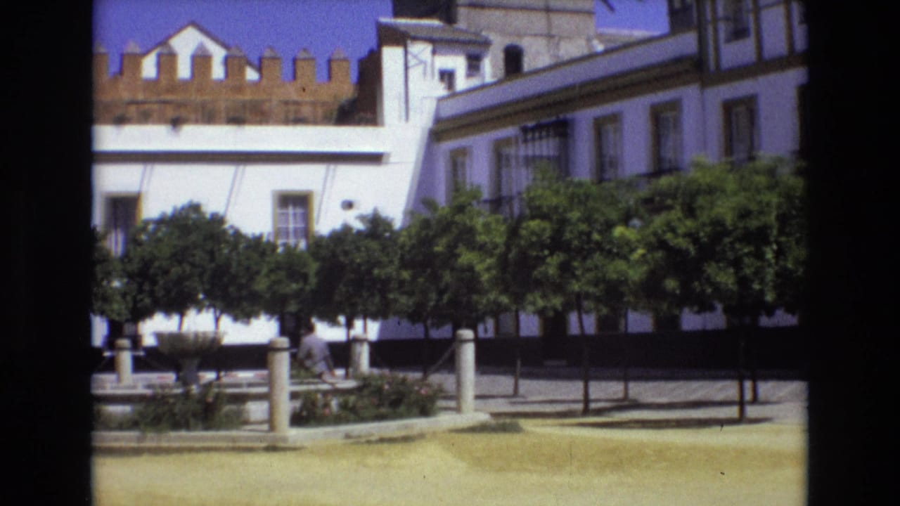 1972 Spain 8mm frame showing a white historic-style building behind trimmed green trees in a bright plaza.