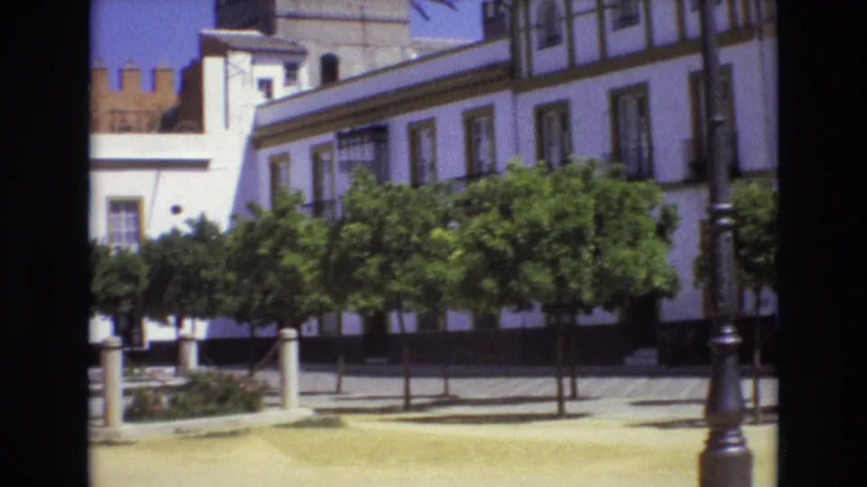 1972 Spain home-movie frame with a white building, green trees, plaza pavement, and a partial ornate lamppost at the right edge.