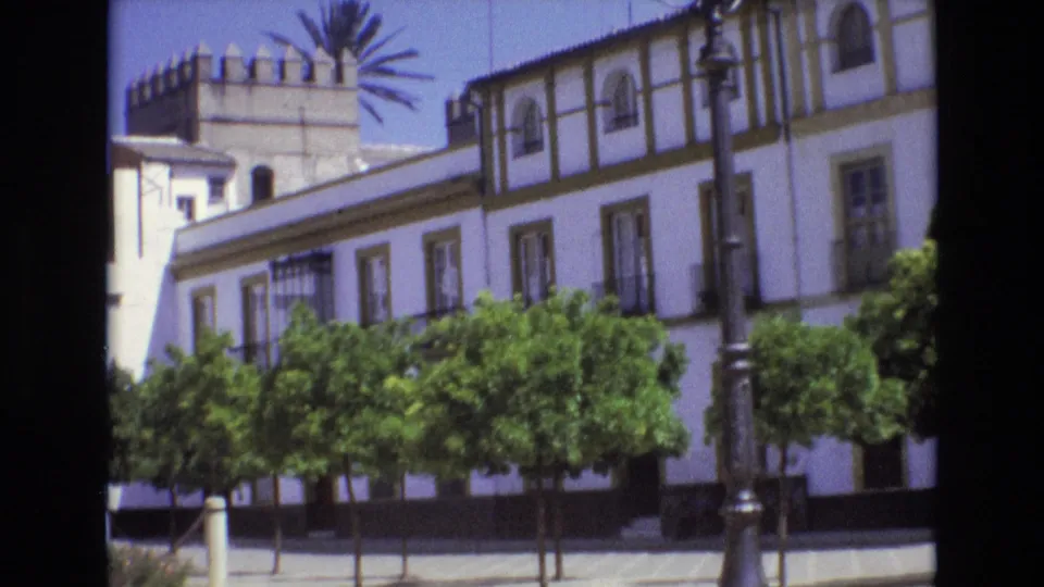 1972 Spain 8mm frame showing a closer tilted view of the white building facade, trees, palm fronds, and an ornate lamppost.