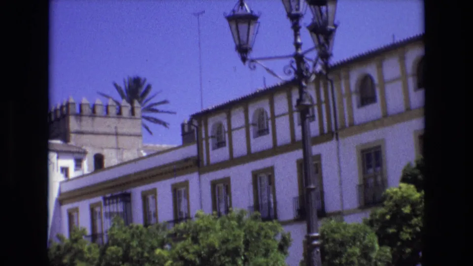 1972 Spain home-movie frame with an ornate streetlamp foregrounding a white historic-style building, palm crown, and green plaza trees.