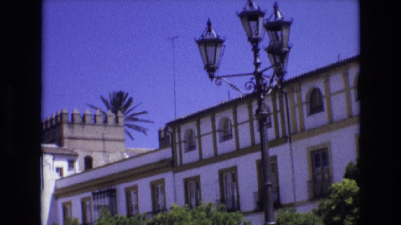 1972 Spain 8mm frame showing a white historic-looking building with a crenellated watchtower, arched windows, ornate black streetlamps, and clear blue sky.