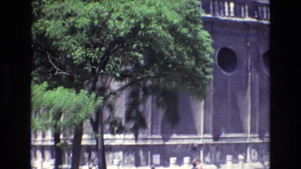 1972 Spain 8mm frame showing a grey stone architectural facade partly obscured by a leafy green tree, with a circular window and decorative upper stonework.