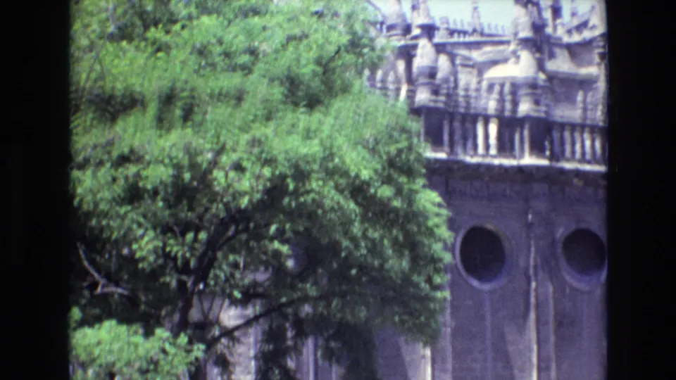 1972 Spain 8mm frame showing dense green foliage in front of an ornate grey stone building with circular windows, carved trim, and decorative upper architecture.