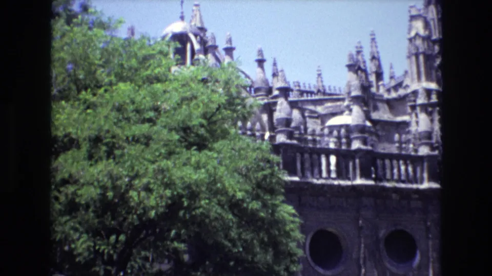 1972 Spain 8mm frame showing an ornate grey stone structure with spires, rounded openings, circular windows, and leafy trees across the foreground.