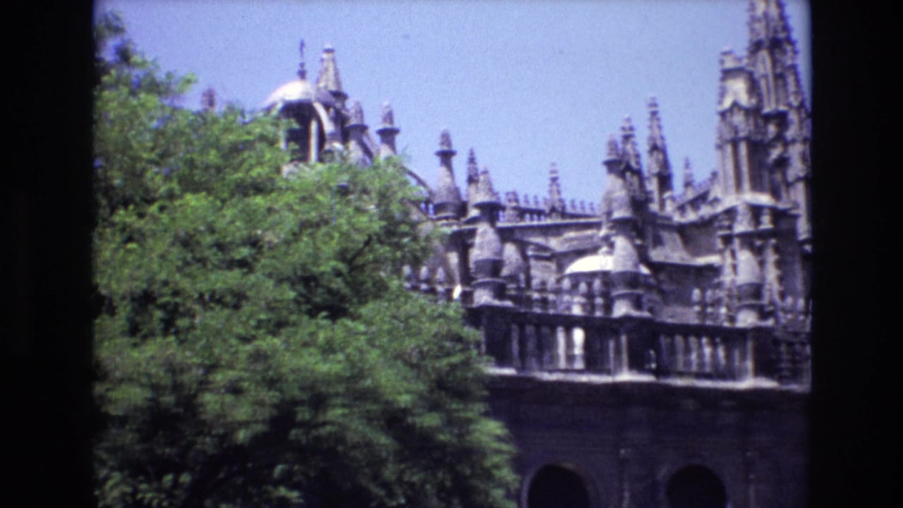 1972 Spain home-movie frame showing an ornate Gothic-style stone building with pinnacles, domed details, and leafy trees in the foreground.