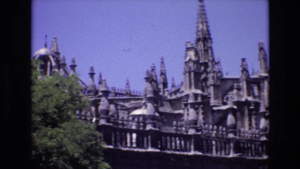 1972 Spain travel footage frame with Gothic-style pinnacles, carved stonework, a balustrade, and trees partially covering the facade.