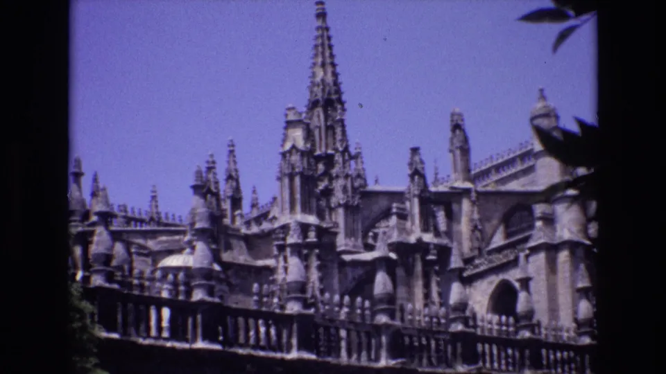 1972 Spain 8mm frame showing a Gothic-style stone facade with a tall spire, arched opening, balustrade, and dark foreground leaves.