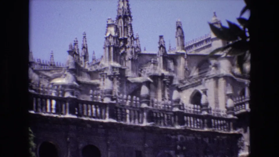 1972 Spain home-movie frame of an ornate Gothic-style exterior with spires, carved roof details, arched openings, and leafy branches at the right edge.
