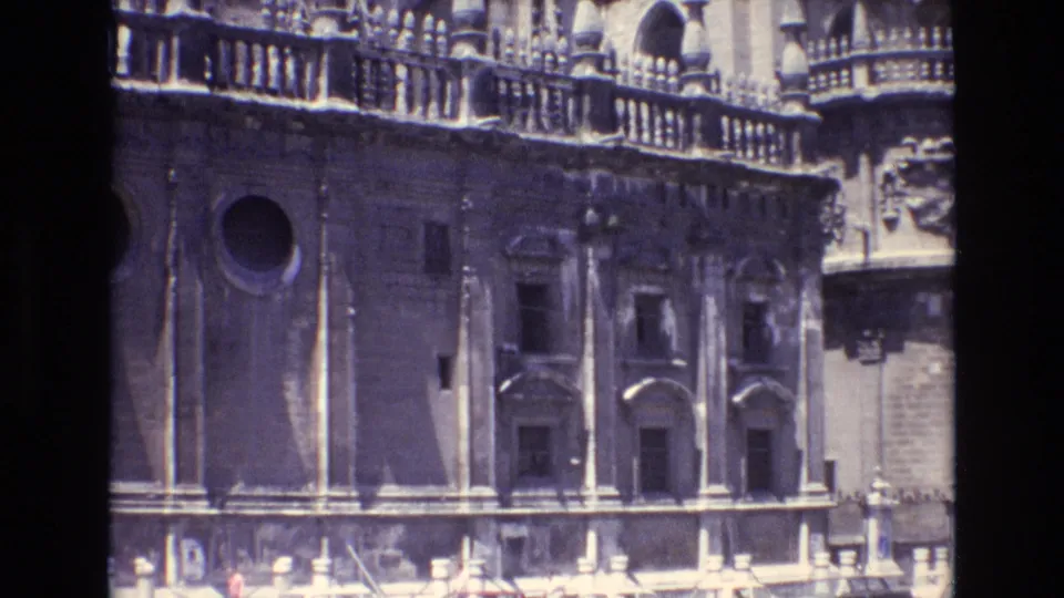 1972 Spain home-movie still showing the lower cathedral facade with a circular window, arched door details, stone balustrade above, and dark projection borders.