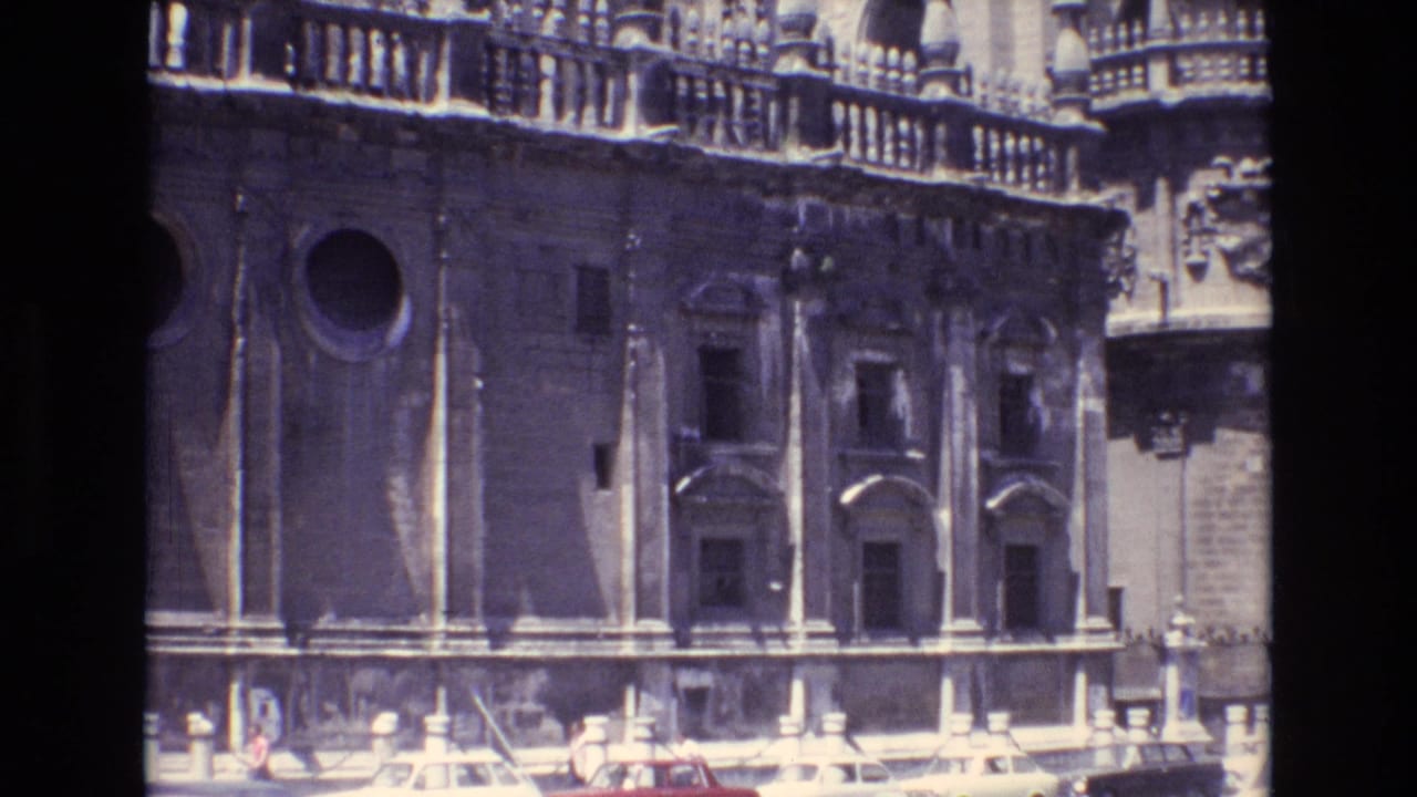 1972 Spain 8mm frame showing a sunlit historic stone facade with carved details, arched windows, a round opening, and partial street-level figures at the bottom edge.