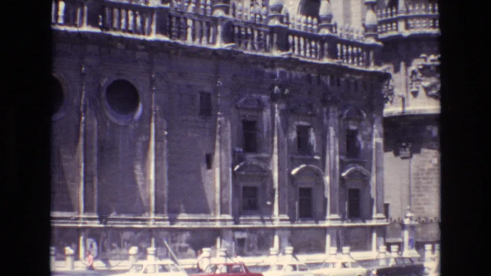 1972 Spain 8mm frame showing a sunlit historic stone facade with carved details, arched windows, a round opening, and partial street-level figures at the bottom edge.