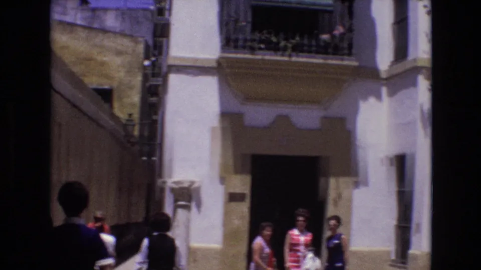 1972 Spain 8mm frame showing several pedestrians or tourists on a sunlit narrow street beside a whitewashed building with wrought-iron balcony railings and a shaded entrance.