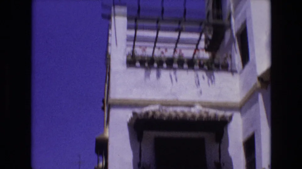 1972 Spain 8mm frame showing a low-angle look at a whitewashed building facade with a projecting balcony, dark iron railing, roofline detail, and clear blue sky.