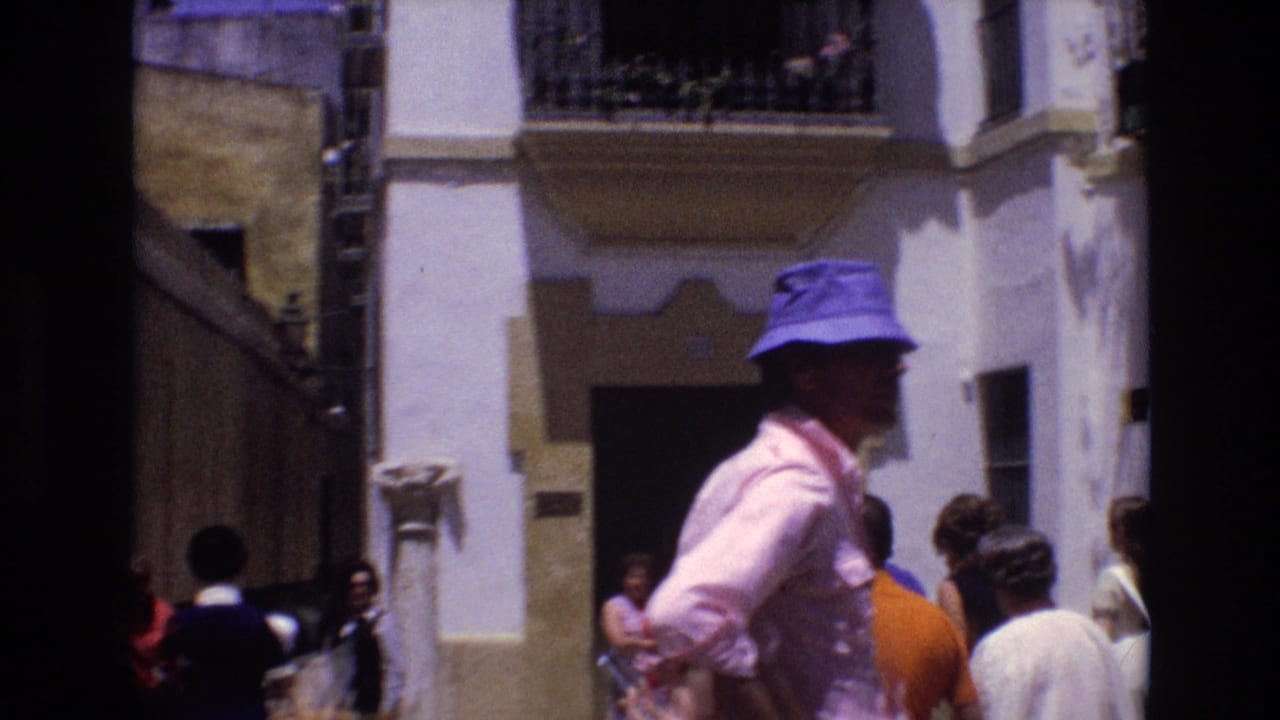 In Spain 1972 home-movie footage, a man in a blue bucket hat and pink shirt stands in the foreground as pedestrians gather along a narrow street with white buildings and balconies.