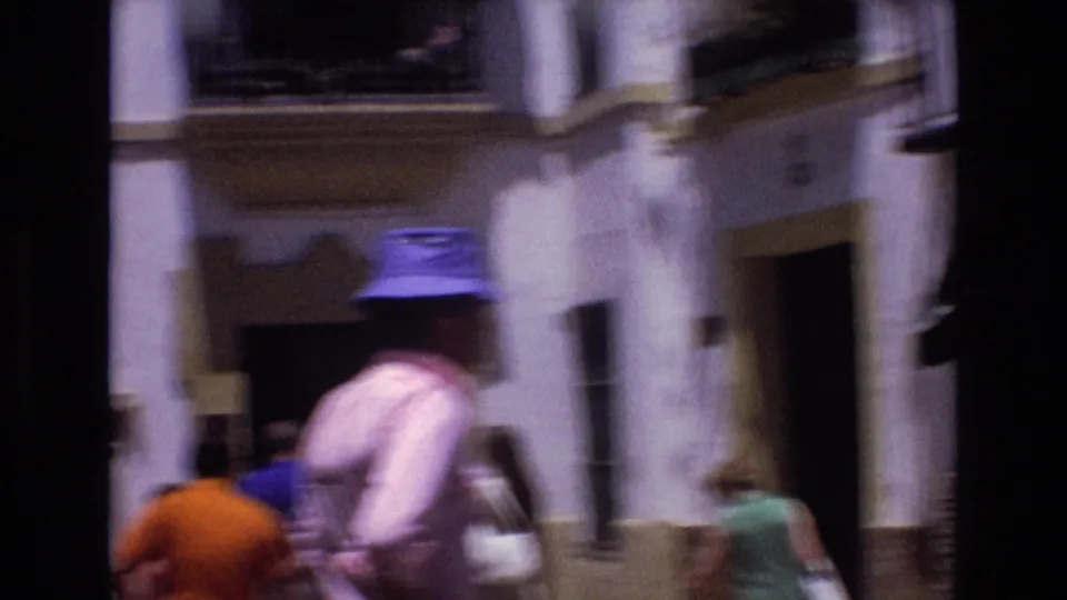 The man in the blue hat appears motion-blurred in the foreground while people move through a bright Spanish street lined with white facades and dark window openings.