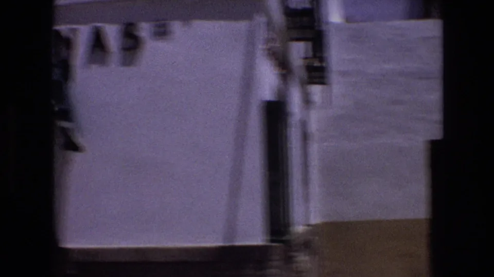 A handheld camera movement catches a soft, blurred view of a white stucco wall, doorway, and edge of the narrow street, with only partial figures at the margins.