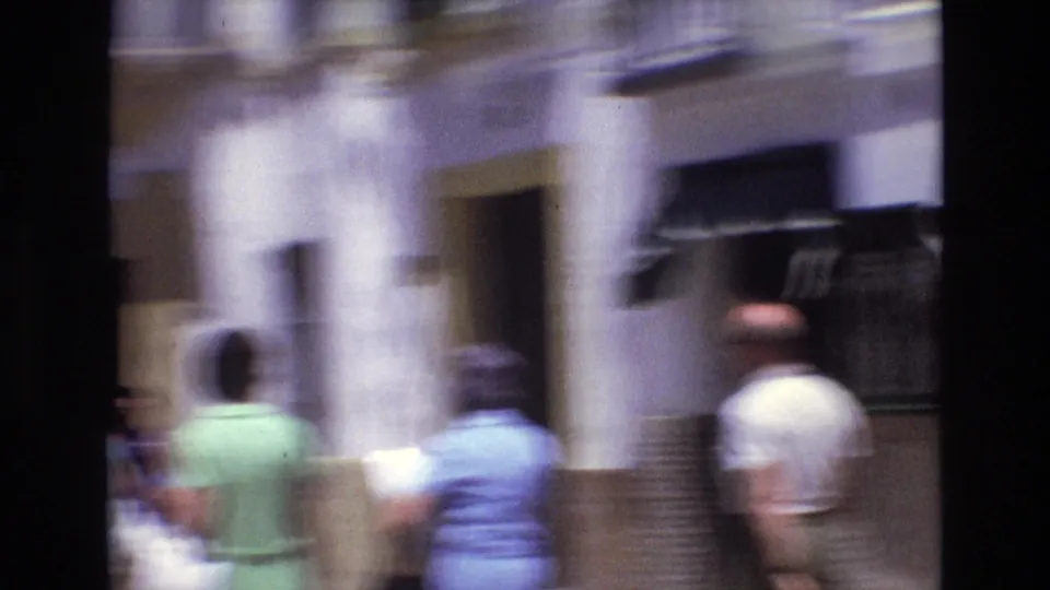 Several pedestrians in casual clothing walk away from the camera along a narrow Spanish street with white facades, shuttered openings, and strong sunlit contrast.