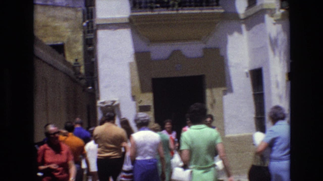 1972 Spain home-movie frame showing people in summer clothing moving through a narrow street beside a white stucco facade with a dark doorway.