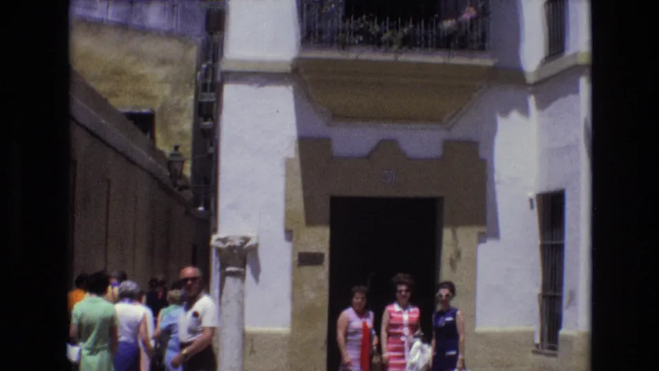 1972 Spain street frame with small groups of pedestrians near a dark doorway, barred window, and white stucco wall in bright sunlight.