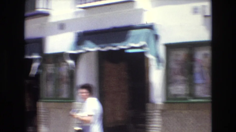 Soft, motion-blurred 1972 Spain home-movie frame showing a man in a light shirt passing a building frontage with windows and an awning-like edge.