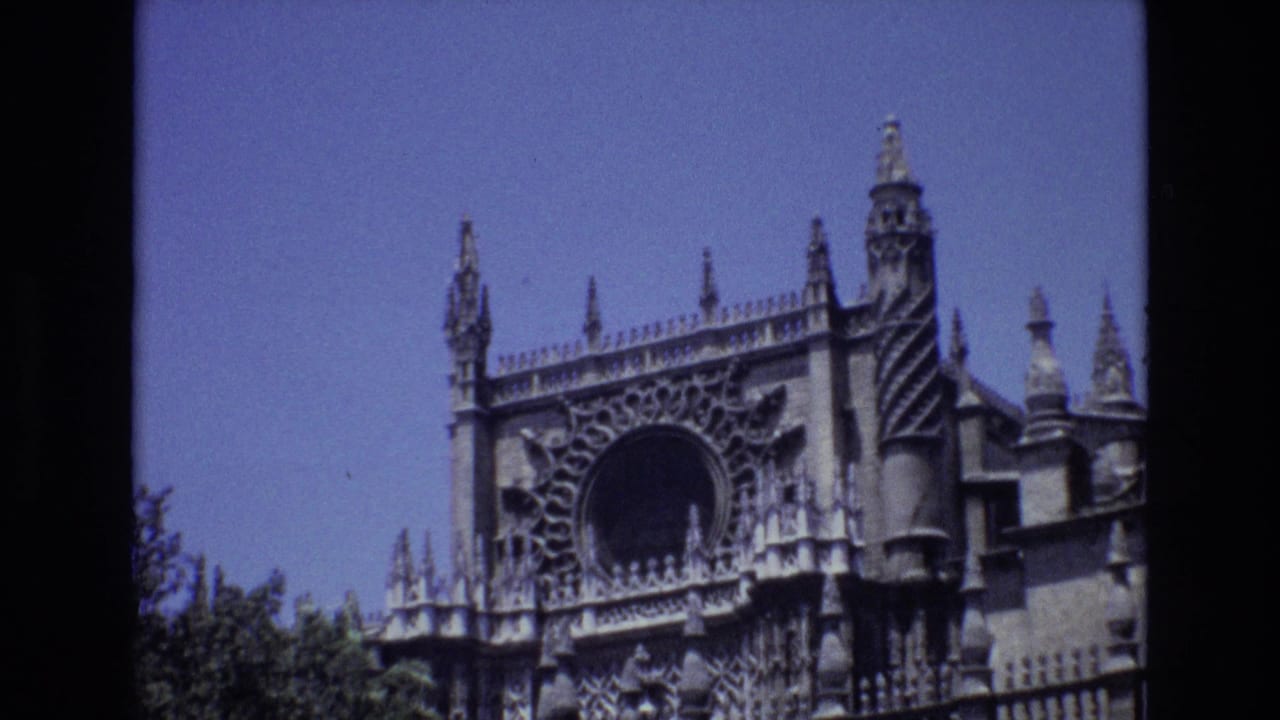 1972 Spain 8mm frame showing an ornate Gothic-style stone facade with spires, carved tracery, a large circular opening, blue sky, and trees below.