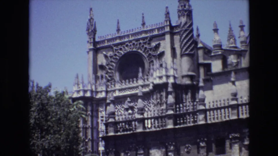 1972 Spain 8mm frame with the camera centered on the upper portion of an elaborate historic stone building, showing spires, decorative tracery, a circular opening, and tree canopy.