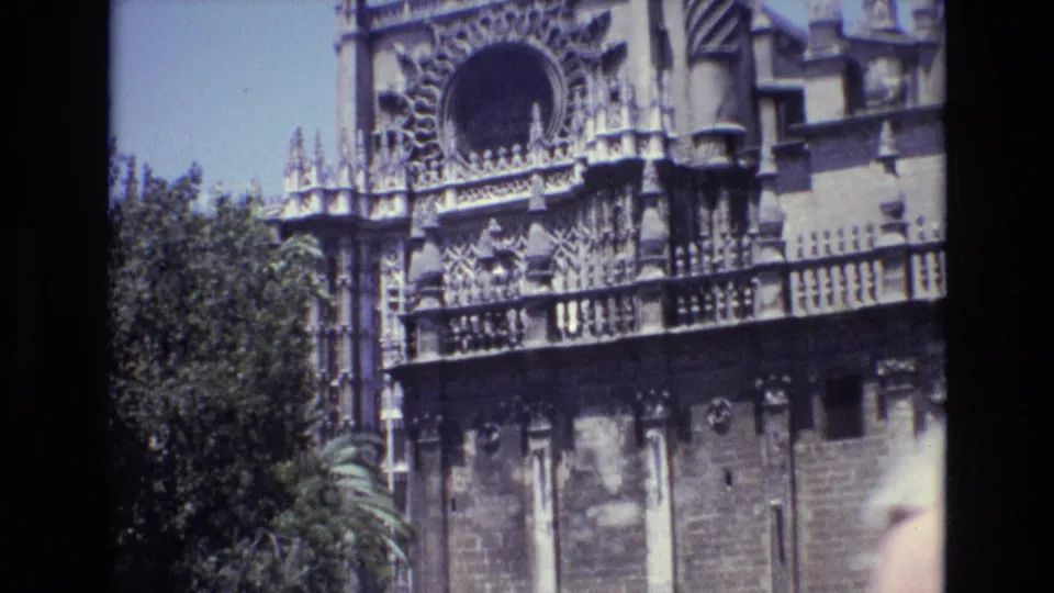 1972 Spain 8mm frame showing a visitor-level view of an ornate historic facade with spires, carved stonework, leafy trees, and lower masonry panels.