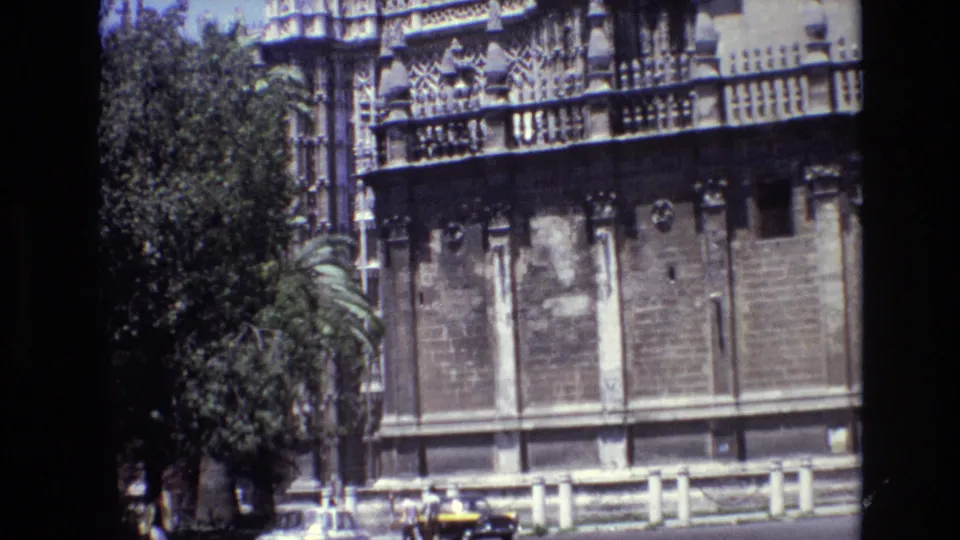 1972 Spain 8mm frame showing the historic stone building from upper ornament down to the street edge, with trees, lower wall panels, and a small yellow car visible near the bottom.