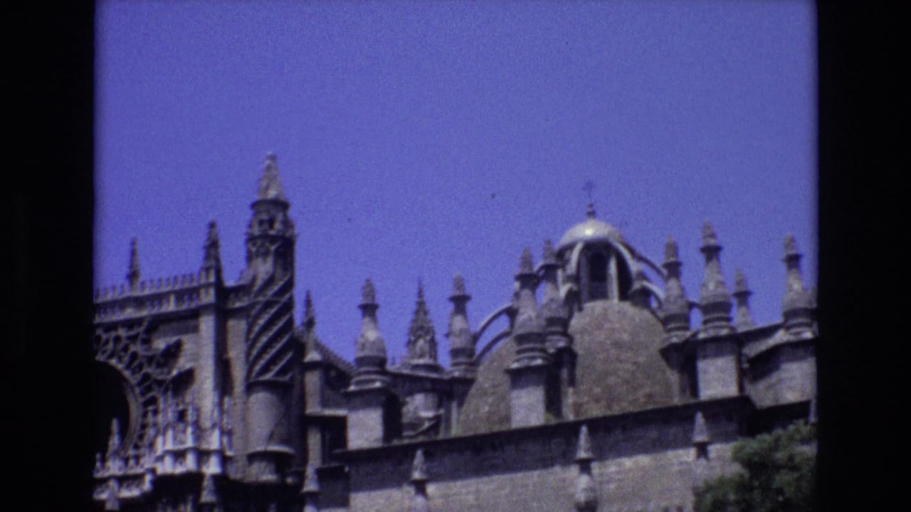 1972 Spain 8mm frame showing the upper facade of a Gothic church with stone spires, decorative carving, and a domed roof feature against a clear blue sky.
