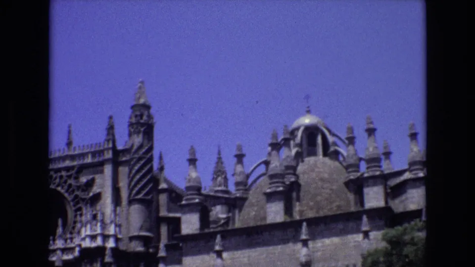 1972 Spain 8mm frame showing the upper facade of a Gothic church with stone spires, decorative carving, and a domed roof feature against a clear blue sky.